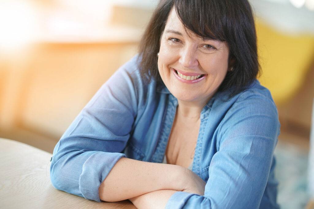 Hymyilevä keski-ikäinen nainen kotona – hyvinvointia ja tasapainoa estrogeenin vähenemisen jälkeen.